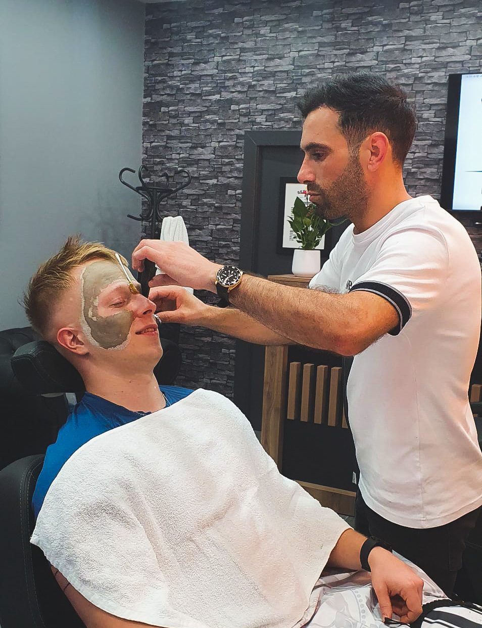 Serhan applying a face mask to a client reclined in the barber’s chair